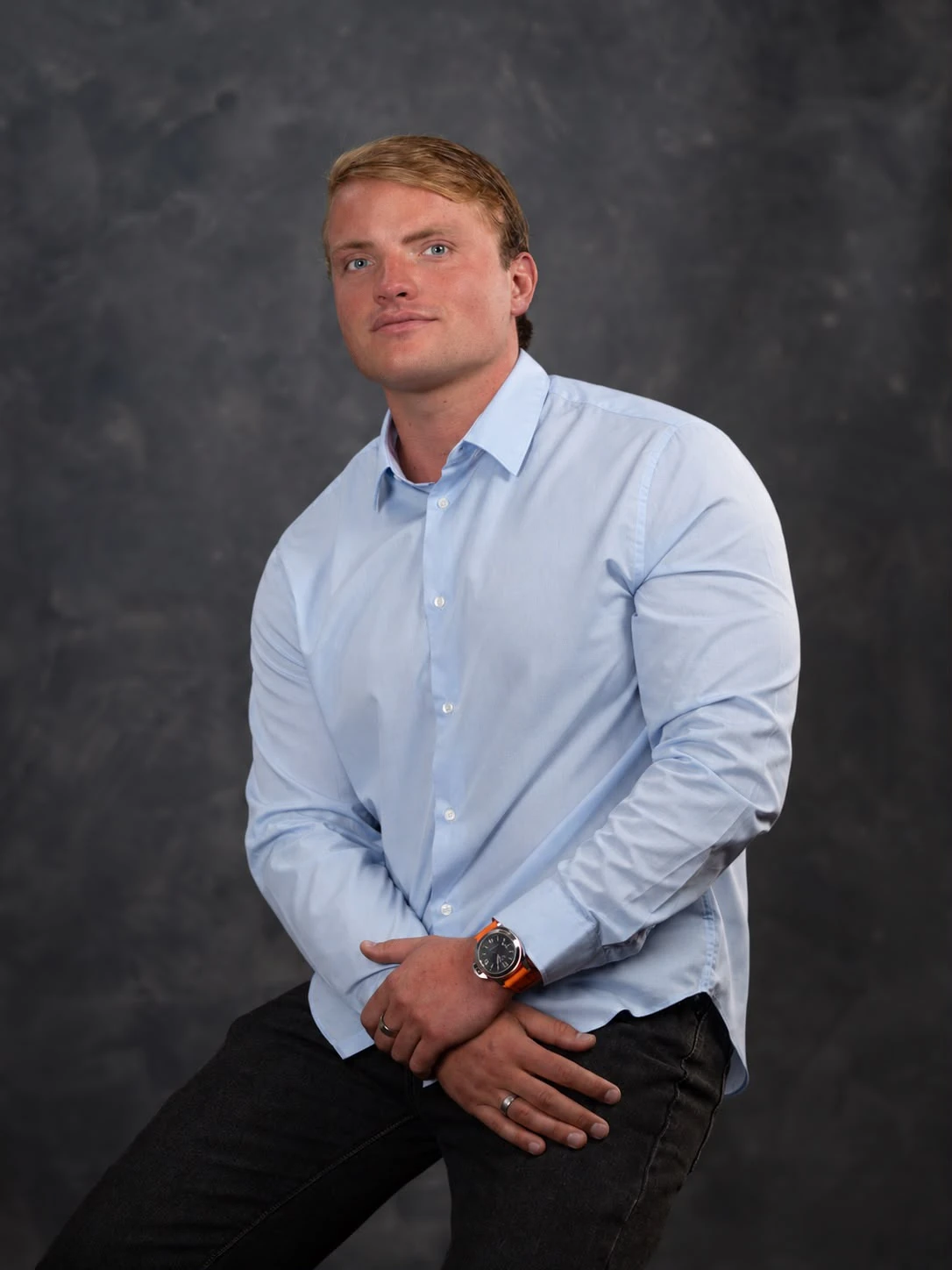 Oliver de Bruin seated in a light blue shirt against a charcoal studio background